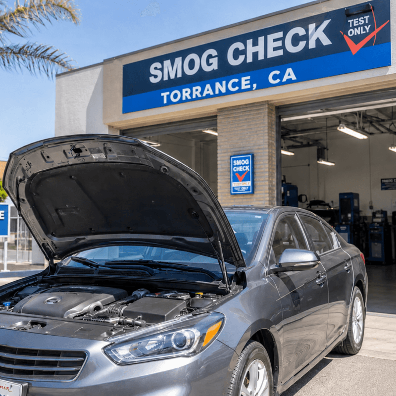 A calm driver confidently pulling into a professional smog testing station in Torrance, California. Show the moment of arrival with the testing facility visible and welcoming. Include subtle visual elements of vehicle maintenance (open hood showing clean engine, fuel cap being checked).